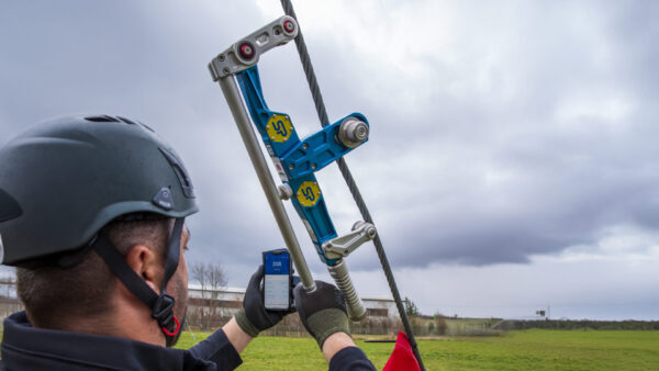 Technician inspecting cable device outdoors with smartphone and safety gear under cloudy skies.